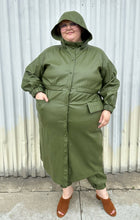 Load image into Gallery viewer, Full-body front view of a size 22/24 Eloquii olive green pleather trench coat and pants two-piece set styled buttoned up with the hood up a black tee on a size 22/24 model. The photo is taken outside in natural lighting.
