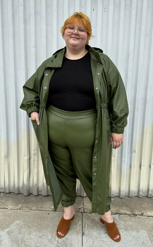 Full-body front view of a size 22/24 Eloquii olive green pleather trench coat and pants two-piece set styled with a black tee on a size 22/24 model. The photo is taken outside in natural lighting.