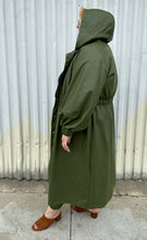 Load image into Gallery viewer, Full-body side view of a size 22/24 Eloquii olive green pleather trench coat and pants two-piece set styled with the hood up a black tee on a size 22/24 model. The photo is taken outside in natural lighting.
