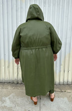 Load image into Gallery viewer, Full-body back view of a size 22/24 Eloquii olive green pleather trench coat and pants two-piece set styled with the hood up a black tee on a size 22/24 model. The photo is taken outside in natural lighting.
