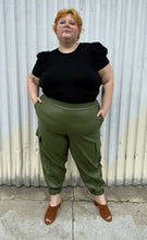 Load image into Gallery viewer, Full-body front view of just the pants of a size 22/24 Eloquii olive green pleather trench coat and pants two-piece set styled with a black tee on a size 22/24 model. The photo is taken outside in natural lighting.
