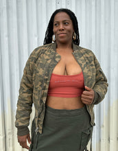 Load image into Gallery viewer, Front view of a size M (fits like 10/12) STAMPD brand camo zip-up bomber jacket styled open over a peach crop and green cargo skirt on a size 14/16 model. The photo is taken outside in natural lighting.
