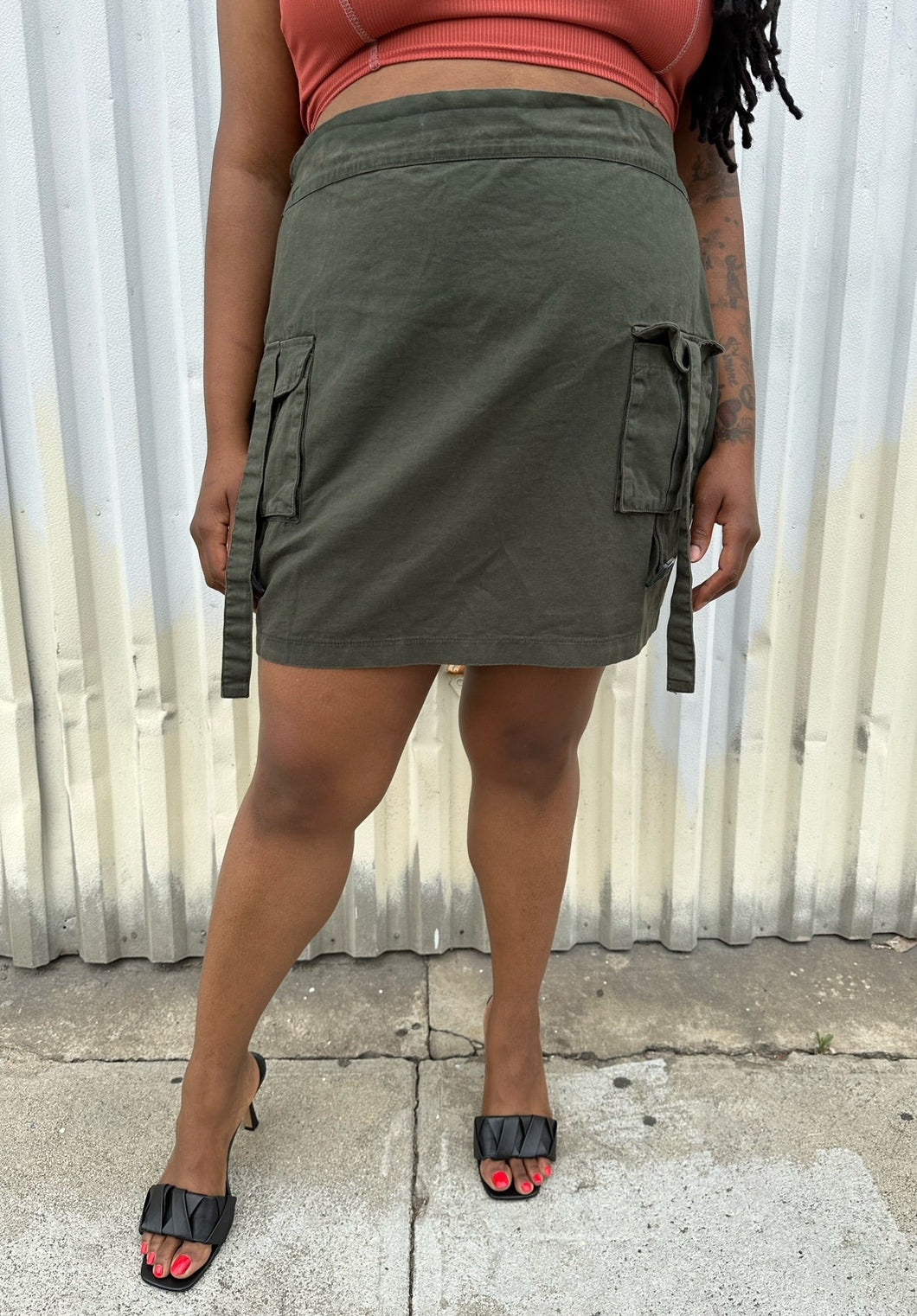 Front view of a size 16 Pretty Little Thing muted army green cargo-style mini skirt with utility strap details styled with a rust-peach crop tank and black kitten heels on a size 14/16 model. The photo is taken outside in natural lighting.