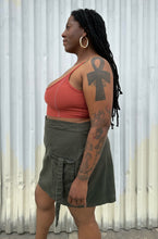 Load image into Gallery viewer, Fuller-body side view of a size 16 Pretty Little Thing muted army green cargo-style mini skirt with utility strap details styled with a rust-peach crop tank and black kitten heels on a size 14/16 model. The photo is taken outside in natural lighting.

