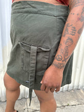 Load image into Gallery viewer, Close up side view of a size 16 Pretty Little Thing muted army green cargo-style mini skirt with utility strap details styled with a rust-peach crop tank and black kitten heels on a size 14/16 model. The photo is taken outside in natural lighting.
