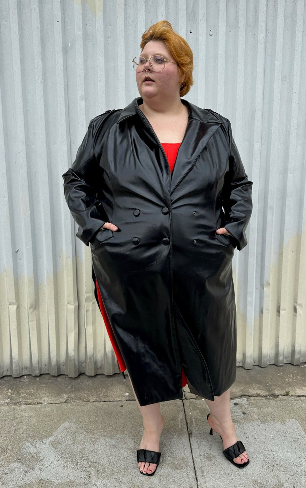Full-body front view of a size 22/24 Eloquii black faux leather double-breasted long coat  with side slits, pockets, and cuffs styled closed over a red dress on a size 22/24 model. The photo was taken outside in natural lighting.