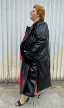 Load image into Gallery viewer, Full-body side view of a size 22/24 Eloquii black faux leather double-breasted long coat  with side slits, pockets, and cuffs styled open over a red dress on a size 22/24 model. The photo was taken outside in natural lighting.
