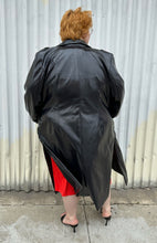 Load image into Gallery viewer, Full-body back view of a size 22/24 Eloquii black faux leather double-breasted long coat  with side slits, pockets, and cuffs styled open over a red dress on a size 22/24 model. The photo was taken outside in natural lighting.
