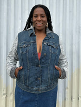 Load image into Gallery viewer, Front view of a size 1 Torrid medium wash denim jacket with hoodie sleeves and hood buttoned up over a rust-peach crop tank and denim skirt on a size 14/16 model. The photo is taken outside in natural lighting.
