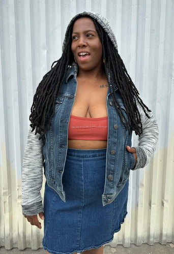 Front view of a size 1 Torrid medium wash denim jacket with hoodie sleeves and hood styled open and with the hood up over a rust-peach crop tank and denim skirt on a size 14/16 model. The photo is taken outside in natural lighting.