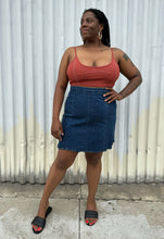 Load image into Gallery viewer, Full-body front view of a size 16 ASOS medium wash denim pencil skirt styled with a rust-peach cropped tank and black flats on a size 14/16 model. The photo is taken outside in natural lighting.
