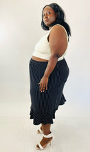 Load image into Gallery viewer, A full-body side view of a size 4X Maggie Barnes black maxi skirt with white pinstripes and a ruffle hem skirt styled with a white crop tank and white strappy heels on a size 24/26.
