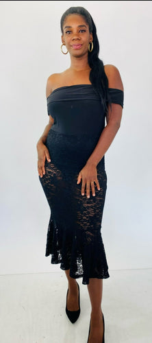 Full-body front view of this Fashion to Figure size 1 off-the-shoulder black bodysuit maxi dress with black lace overlay and mermaid-style skirt styled with black heels on a size 12 model.
