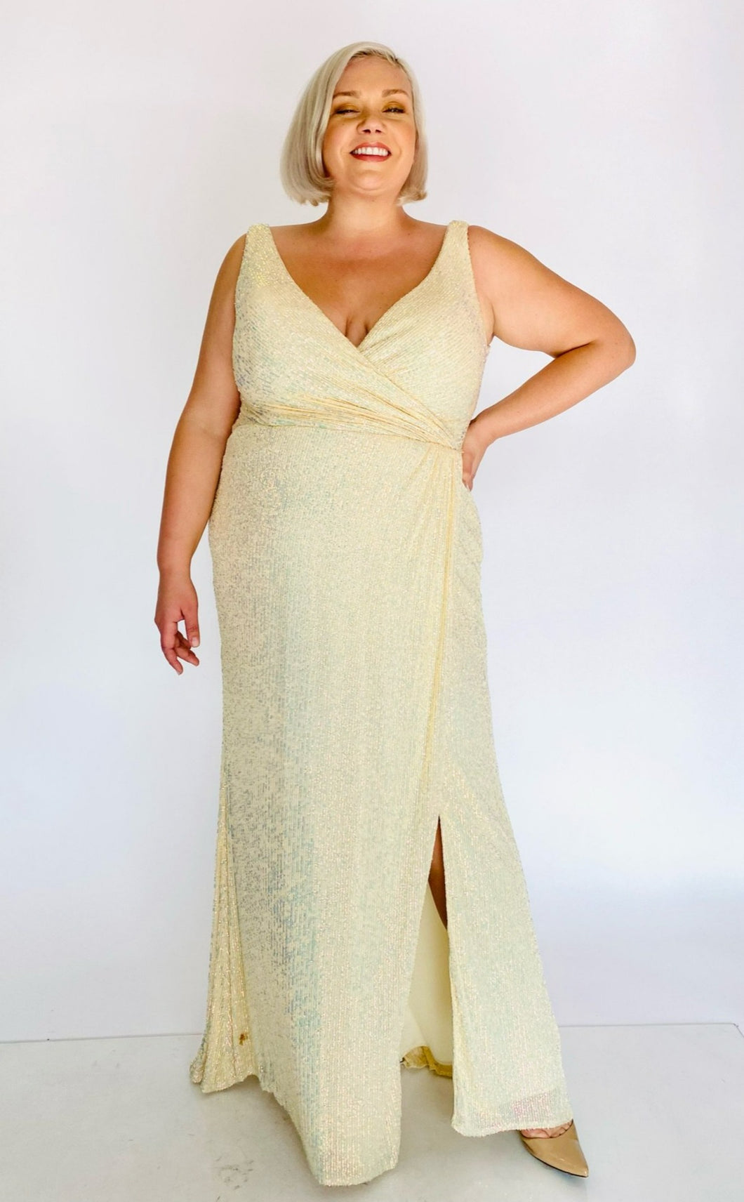 Full-body front view of a size 20 La Femme Curve shimmery champagne yellow gown with a faux wrap bust and high side slit styled with tan pumps on a size 18/20 model.