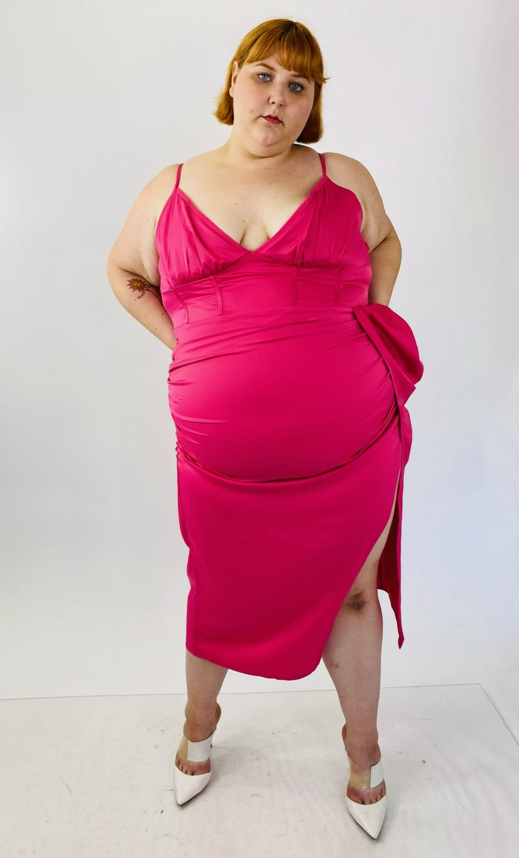 Full-body front view of a size 22 Pretty Little Thing hot pink strappy midi dress with a high side slit and structured boning at the bust styled with white heels on a size 22 model.