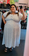 Load image into Gallery viewer, Additional full-body front view of a size 3X Who What Wear baby blue and white all-over floral print maxi dress with elastic waist, subtle puff sleeves, and a tie bust detail styled with gray feather earrings and black heels on a size 22/24 model.
