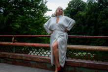 Load image into Gallery viewer, Full-body front view of a Monique Lhuillier for 11Honoré silky white gown with black mini butterfly print, choker and kethole details, and puff sleeves on a size 18/20 model standing on a bridge by a river.
