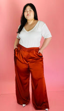 Load image into Gallery viewer, Full-body front view of a pair of size 16 Jonathan Simkhai rust-colored shiny, silky pants with an elastic waist, wide legs, and a lacy side slit detail styled with a white tee on a size 14/16 model.
