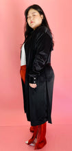 Load image into Gallery viewer, Full-body side view of a size 3 Baja East for 11 Honoré black satin duster trench with a belt styled open over a white tee and rust orange silky pants on a size 14/16 model.
