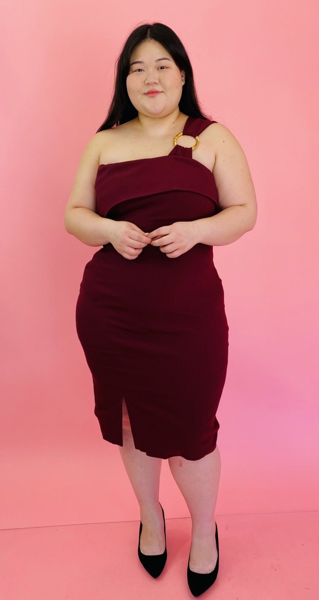 Full-body front view of a size 18 Haney for 11 Honoré burgundy one-shoulder bodycon midi dress with a fold-over bust detail, circle ring strap detail, and slit at the front styled with black pumps on a size 14/16 model.