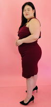 Load image into Gallery viewer, Full-body side view of a size 18 Haney for 11 Honoré burgundy one-shoulder bodycon midi dress with a fold-over bust detail, circle ring strap detail, and slit at the front styled with black pumps on a size 14/16 model.
