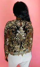 Load image into Gallery viewer, Back view of a size L Naeem Khan for 11 Honoré black long sleeve mesh semi-sheer blouse with intricate gold sequin pattern and details all throughout and sheer sleeves, styled with white trousers on a size 10/12 model.
