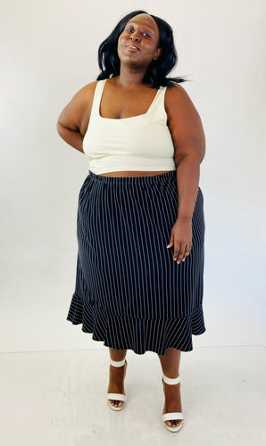 An additional full-body front view of a size 4X Maggie Barnes black maxi skirt with white pinstripes and a ruffle hem skirt styled with a white crop tank and white strappy heels on a size 24/26.