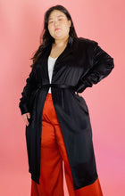 Load image into Gallery viewer, Front view of a size 3 Baja East for 11 Honoré black satin duster trench with a belt styled closed over a white tee and rust orange silky pants on a size 14/16 model.
