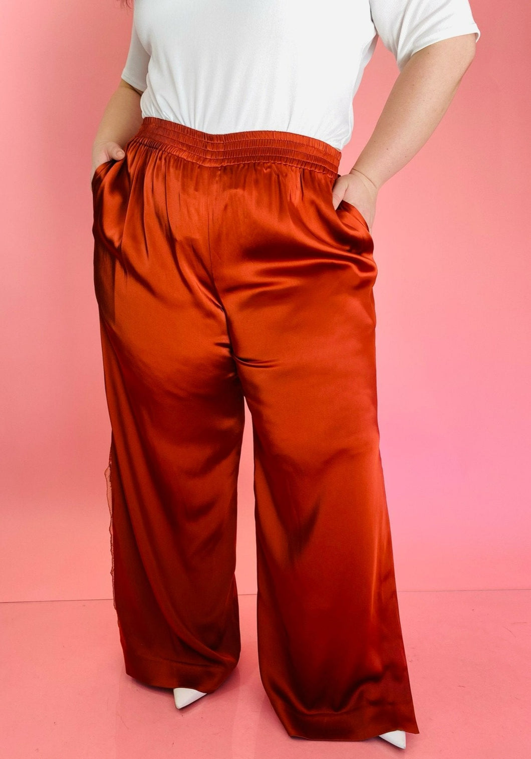 Front view of a pair of size 16 Jonathan Simkhai rust-colored shiny, silky pants with an elastic waist, wide legs, and a lacy side slit detail styled with a white tee on a size 14/16 model.