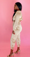 Load image into Gallery viewer, Full-body side view of a size 12 Haney for 11 Honoré gold, sand color collared button-up jumpsuit with belt and tapered, crop ankle detail styled with tan pumps on a size 12 model. 

