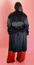 Load image into Gallery viewer, Full-body back view of a size 3 Baja East for 11 Honoré black satin duster trench with a belt styled closed over rust orange silky pants on a size 14/16 model.
