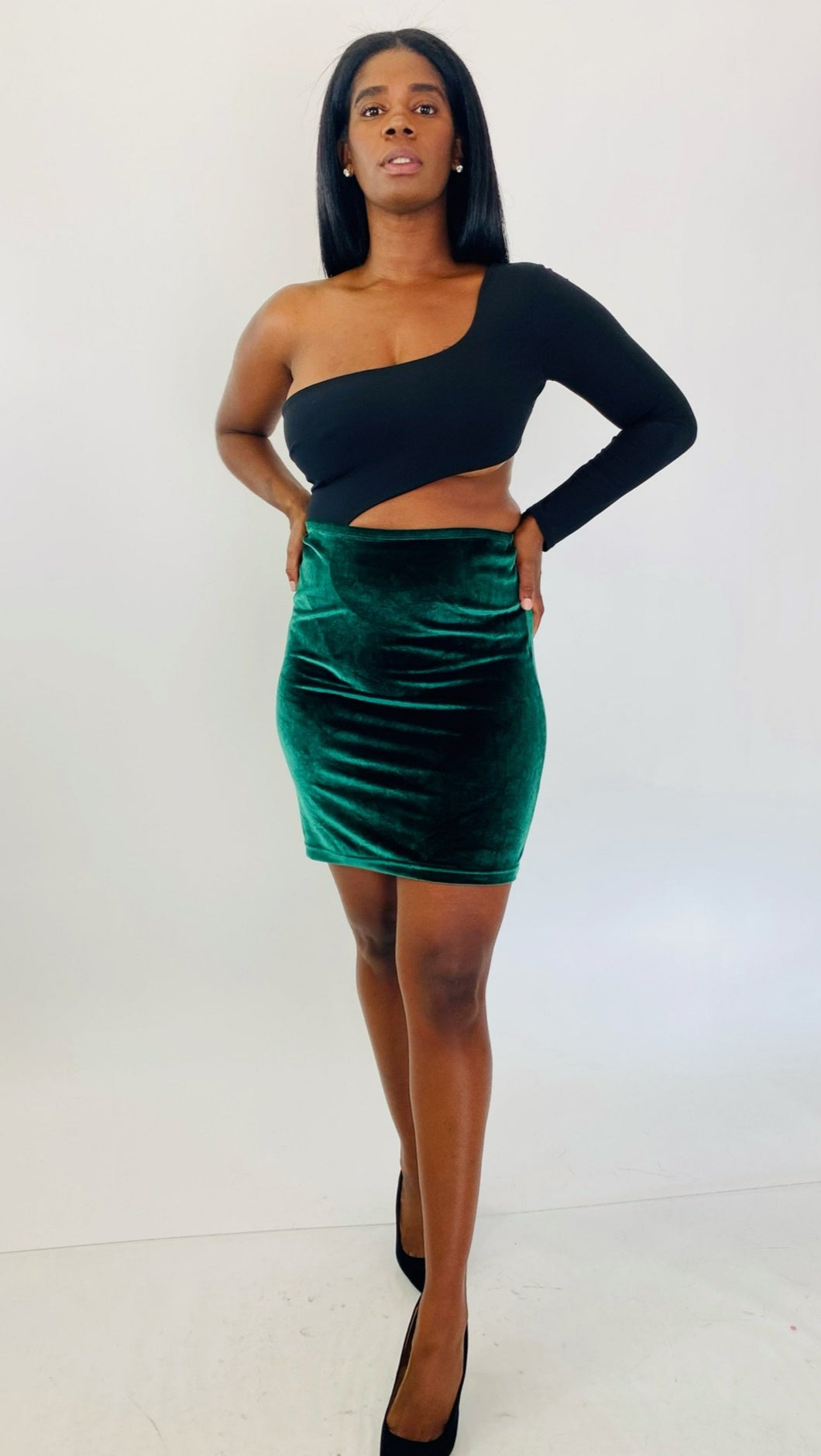 Full-body front view of a size 1X Fashion Nova black one-shoulder long sleeve crop top styled with a green velvet bodycon mini skirt and black pumps on a size 12 model.