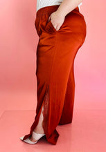 Load image into Gallery viewer, Side view showing off the side slit on a pair of size 16 Jonathan Simkhai rust-colored shiny, silky pants with an elastic waist, wide legs, and a lacy side slit detail styled with a white tee on a size 14/16 model.
