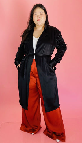 Full-body front view of a size 3 Baja East for 11 Honoré black satin duster trench with a belt styled closed over a white tee and rust orange silky pants on a size 14/16 model.