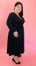 Load image into Gallery viewer, Full-body side view of a size 14 Runway Marc Jacobs x 11 Honoré black long sleeve empire waist midi dress with v-neck faux-wrap bodice and black belt with rosette detail styled with black pumps on a size 14/16 model.
