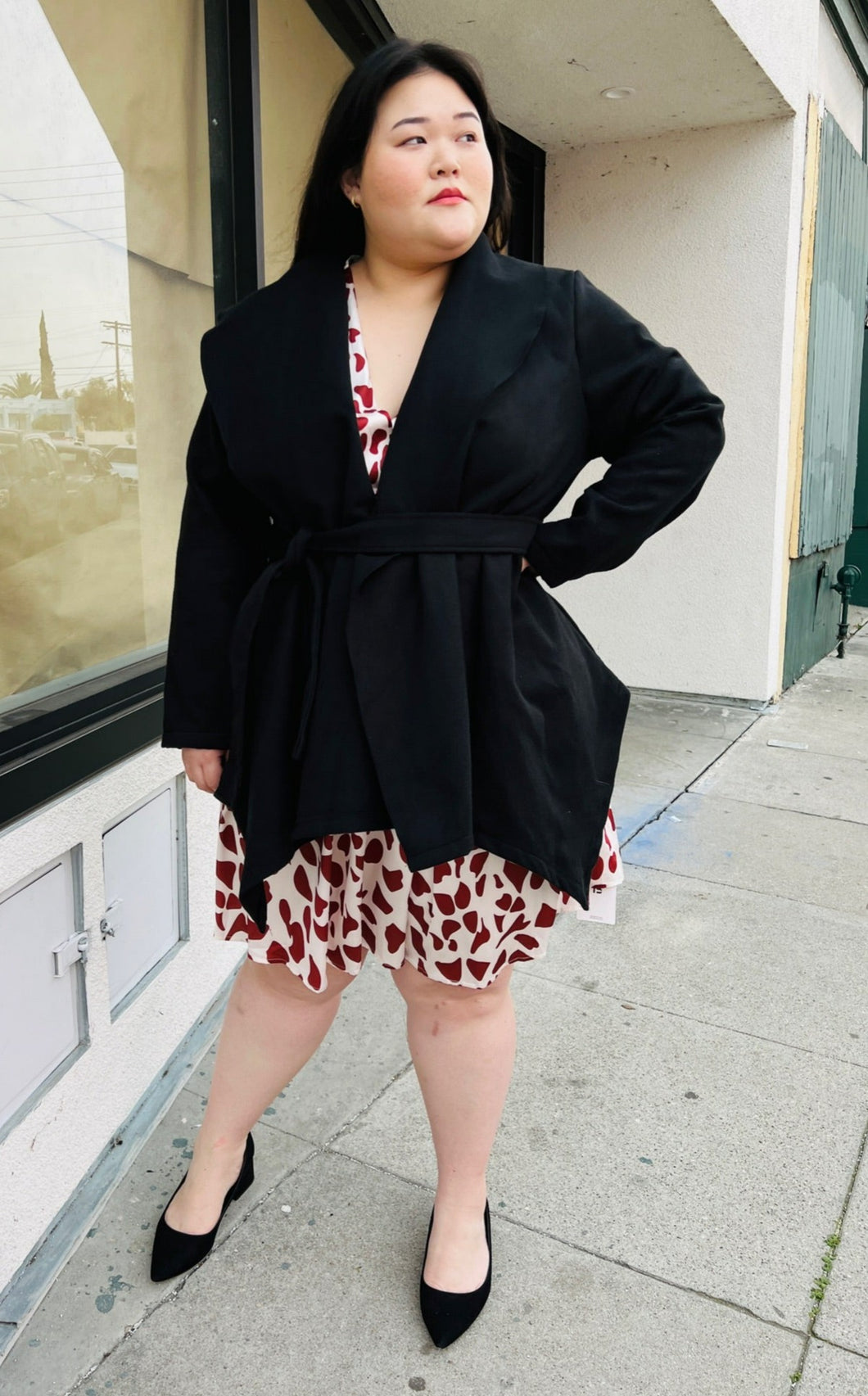 Full-body front view of a Bloomchic black collared coat with a handkerchief hem and a cinching belt styled over a cream and red dress on a size 14/16 model.