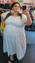 Load image into Gallery viewer, Full-body front view of a size 3X Who What Wear baby blue and white all-over floral print maxi dress with elastic waist, subtle puff sleeves, and a tie bust detail styled with gray feather earrings and black heels on a size 22/24 model.

