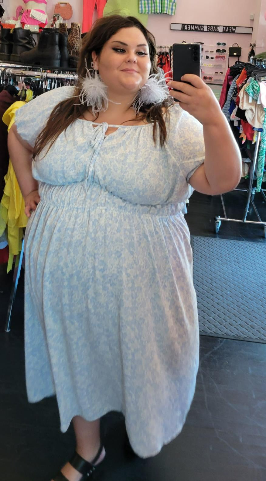 Full-body front view of a size 3X Who What Wear baby blue and white all-over floral print maxi dress with elastic waist, subtle puff sleeves, and a tie bust detail styled with gray feather earrings and black heels on a size 22/24 model.