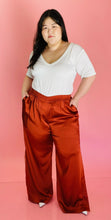 Load image into Gallery viewer, Additional full-body front view of a pair of size 16 Jonathan Simkhai rust-colored shiny, silky pants with an elastic waist, wide legs, and a lacy side slit detail styled with a white tee on a size 14/16 model.
