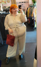 Load image into Gallery viewer, Full-body front view of a size 22 Eloquii off-white/cream all-over sequined button-up jumpsuit with tie belt styled with a maroon faux fur bag and black mary janes on a size 22/24 model.
