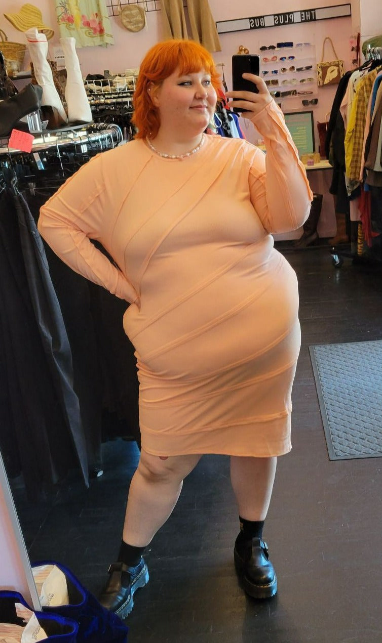 Full-body front view of a size 4 Fashion to Figure pale peach long sleeve ribbed midi dress with textured details styled with black mary janes on a size 22/24 model.