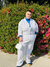 Load image into Gallery viewer, Full-body front view of a size 5X Big Bud Press off-white collared boiler suit/jumpsuit with zip-front details styled over a blue turtleneck and blue boots on a size 22 model in front of a flowering bush.

