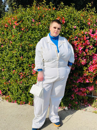 Full-body front view of a size 5X Big Bud Press off-white collared boiler suit/jumpsuit with zip-front details styled over a blue turtleneck and blue boots on a size 22 model in front of a flowering bush.