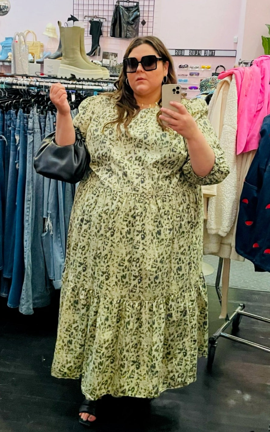 Full-body front view of a sage green tiered maxi dress with darker green leopard spots and puff sleeves styled with black sunglasses, black slides, and a black handbag on a size 22 model.
