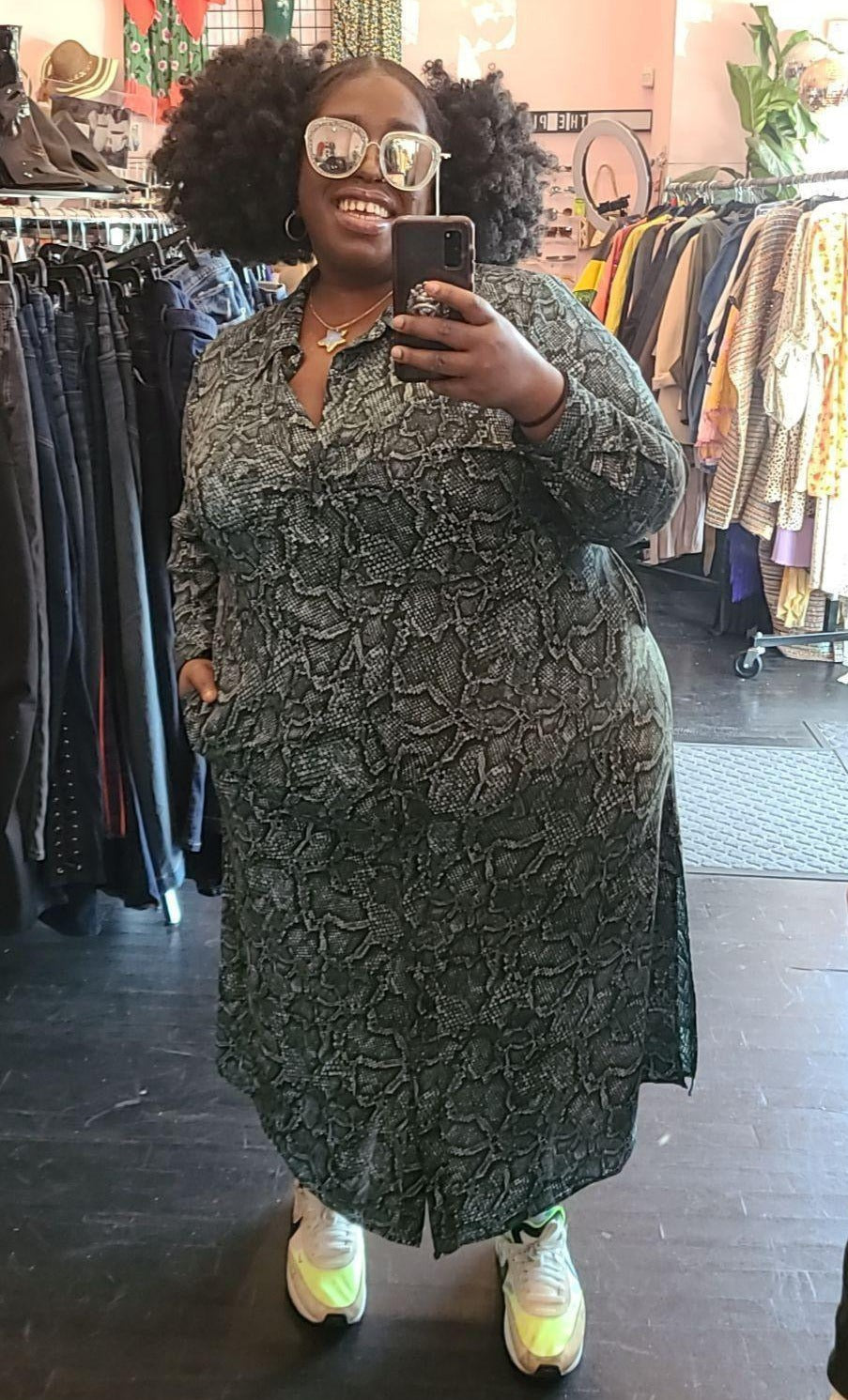 Full-body front view of a size 3X 1. State teal and black snakeprint button-up shirt dress styled with shiny sunglasses and neon sneakers on a size 22/24 model.