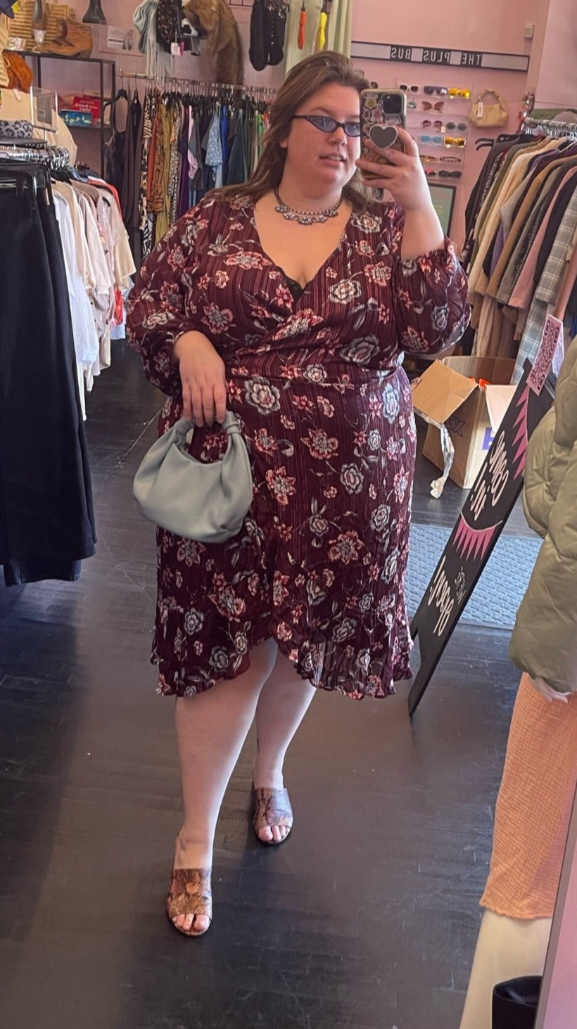 Full-body front view of a size 24 Rachel Roy maroon wrap dress with a ruffled tulip hem, pastel blue and pink floral pattern, silver threading, and long sleeves styled with a pendant, sunnies, and a blue handbag on a size 24 model.