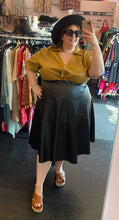 Load image into Gallery viewer, Full-body front view of a size 24 Eloquii camel colored twist-bust detail pleather button-up collared top, styled tucked into a black pleather midi skirt with a black hat on a size 24 model.
