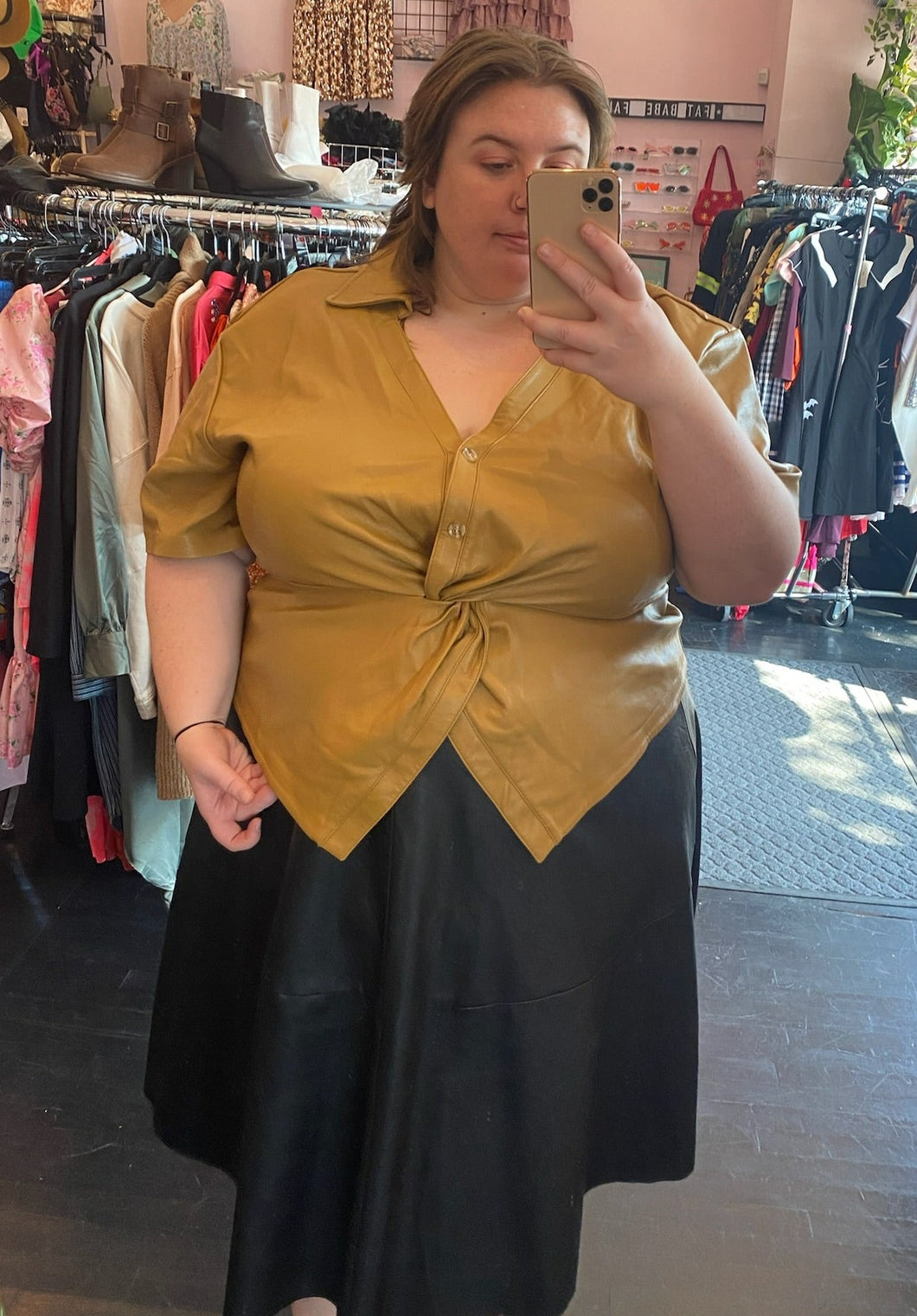 Front view of a size 24 Eloquii camel colored twist-bust detail pleather button-up collared top, styled over a black pleather midi skirt on a size 24 model.