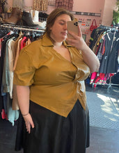Load image into Gallery viewer, Front-side view of a size 24 Eloquii camel colored twist-bust detail pleather button-up collared top, styled over a black pleather midi skirt on a size 24 model.
