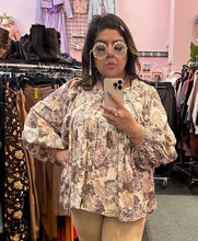 Load image into Gallery viewer, Close up view of a size XXL The Lullaby Club neutral tone floral blouse with fabric buttons, puff sleeves, and a baby doll shape styled with khaki trousers and floral glasses on a size 14/16 model.
